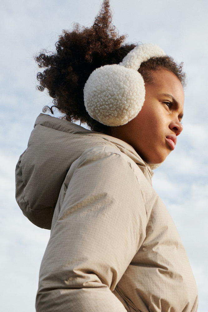 The model wears Liewood Albero Pile Ear Warmers in Sandy, featuring soft recycled fleece, a neutral sandy colour, and a cozy fit. She is wearing a beige zip up jacket and stands against a cloudy sky