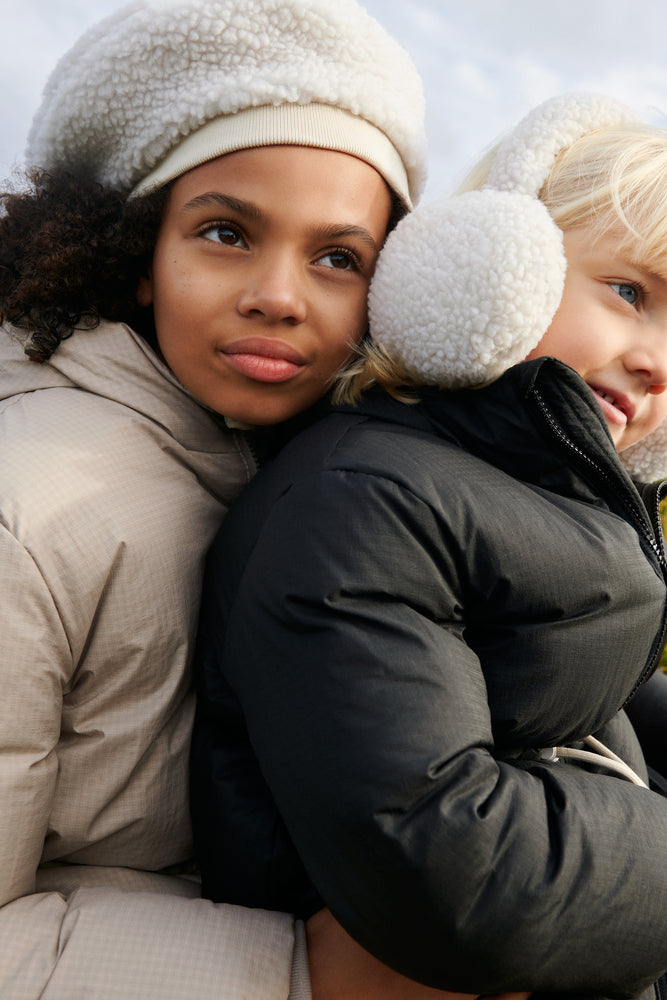 The model wears Liewood Albero Pile Ear Warmers in Sandy, featuring soft recycled fleece, a neutral sandy colour, and a cozy fit. She is wearing a dark green puffa jacket