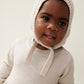 The model wears the Liewood Fresia Baby Hat in Sandy, featuring a playful pompom on top, long adjustable strings, and delicate organic cotton knit