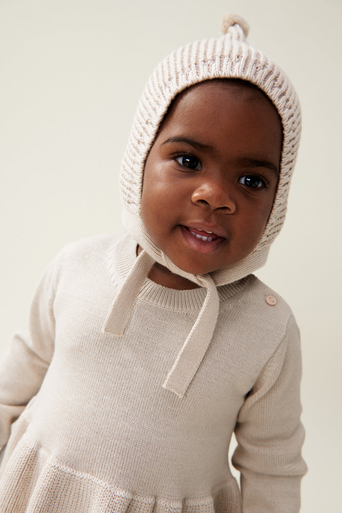 The model wears the Liewood Fresia Baby Hat in Sandy, featuring a playful pompom on top, long adjustable strings, and delicate organic cotton knit