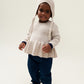 The model wears the Liewood Fresia Baby Hat in Sandy, featuring a playful pompom on top, long adjustable strings, and delicate organic cotton knit, along with a cream frill jumper ad dark trousers