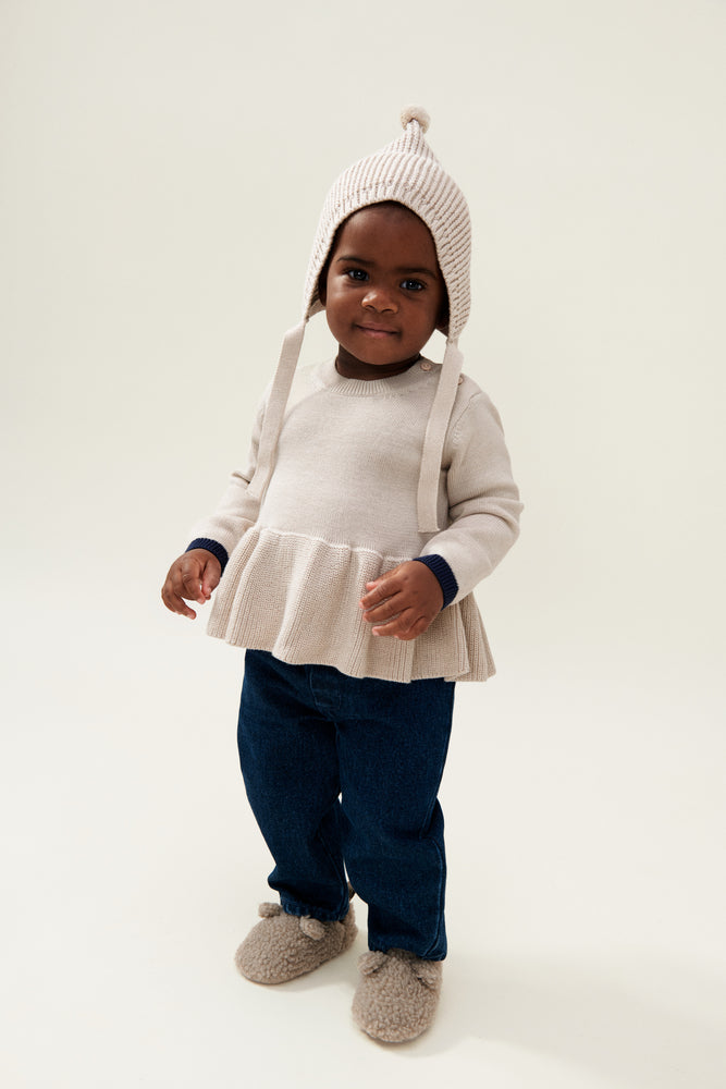 The model wears the Liewood Fresia Baby Hat in Sandy, featuring a playful pompom on top, long adjustable strings, and delicate organic cotton knit, along with a cream frill jumper ad dark trousers