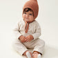 Liewood Tuscany Rose Fresia Baby Hat, featuring a playful pompom on top, long adjustable strings, and delicate organic cotton knit. The baby is sitting and wearing a cream jumpsuit