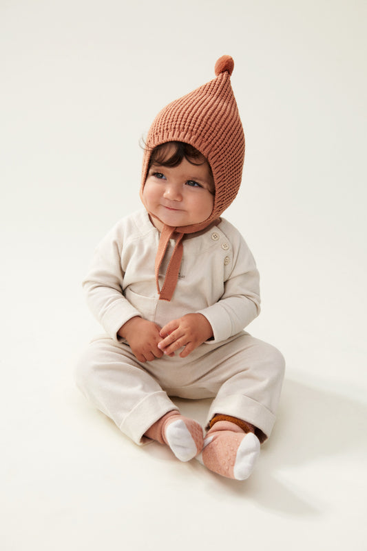 Liewood Tuscany Rose Fresia Baby Hat, featuring a playful pompom on top, long adjustable strings, and delicate organic cotton knit. The baby is sitting and wearing a cream jumpsuit