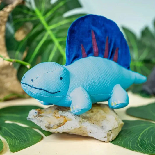 Dimitri the Dimetrodon dinosaur toy perched on a stone among lush green leaves, with a close-up view of its textured vegan leather body and bold navy blue crest