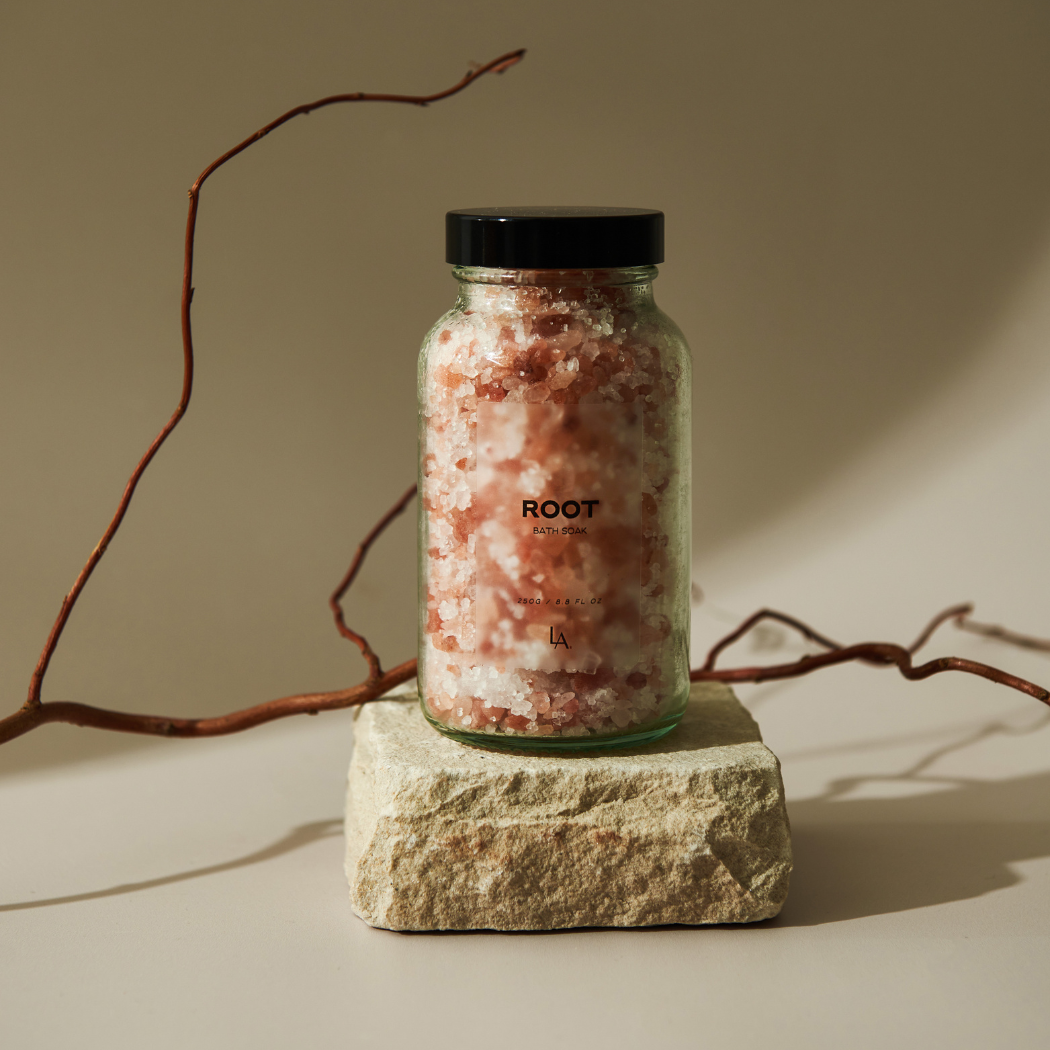 A see through glass jar with a back lid sits on top of a piece of natural stone, with a branch lying behind it. The jar is filled with white and pink bath salts. The jar says ROOT on the front in black writing