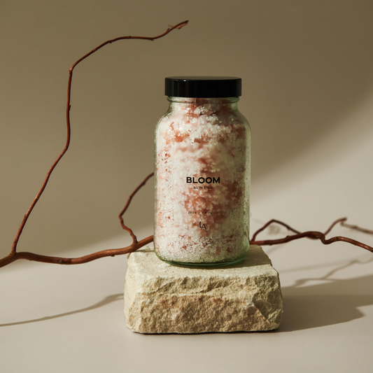 A see through glass jar with a back lid sits on top of a piece of natural stone, with a branch lying behind it. The jar is filled with white and pink bath salts. The jar says BLOOM on the front in black writing