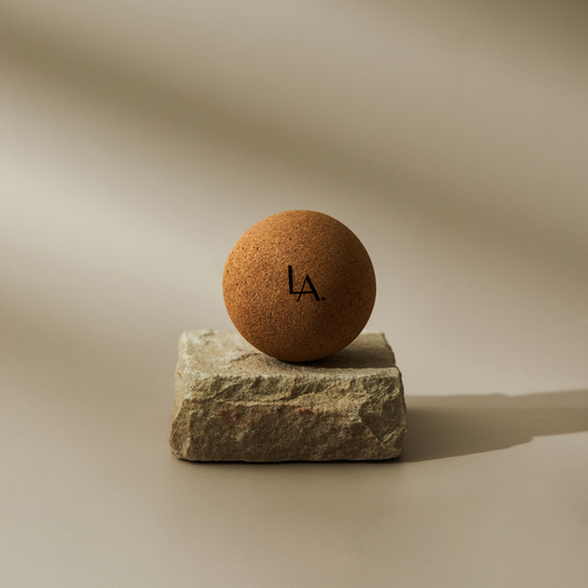 A cork massage balls sits on a slab of natural stone. casting a shadow. The initials LA is printed on it. The background is a simple cream.