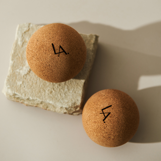 Two cork massage balls sit on a cream background. One rests on a slab of natural stone. They cast shadows. The initials LA are printed on them.