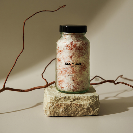 A see through glass jar with a back lid sits on top of a piece of natural stone, with a branch lying behind it. The jar is filled with white and pink bath salts. The jar says SLUMBER on the front in black writing