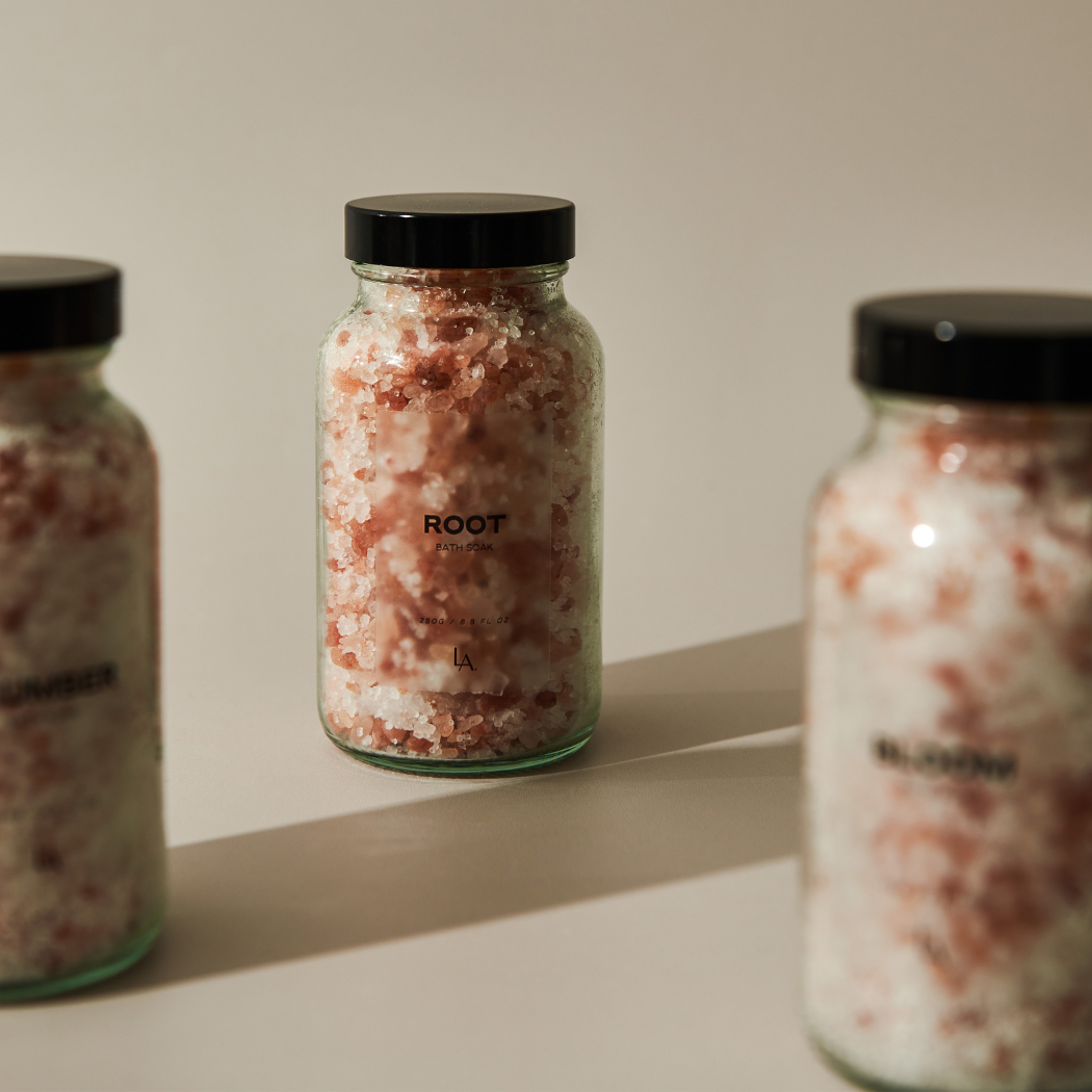 Three clear glass jars with black lids sit on a cream background casting shadows. They contain white and pink bath salts.