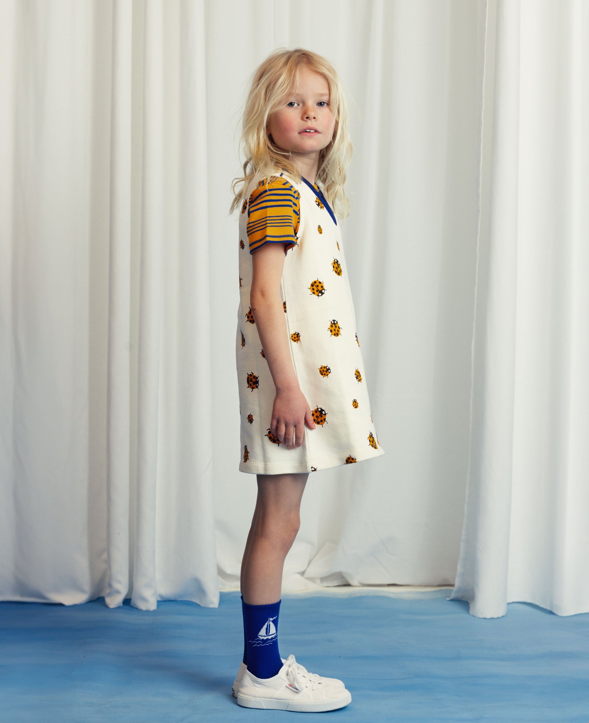 A young girl with blonde hair wears a short-sleeved dress featuring a floral print on an off-white base, layered over a striped T-shirt. She pairs the outfit with bright blue socks with a sailboat design and white trainers, standing against a white curtain backdrop.
