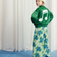 A young child wearing a green satin bomber jacket with an oversized white embroidered music note on the back, paired with wide-leg trousers in a blue floral print.