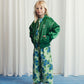 A child styled in a green bomber jacket with piping details, blue floral wide-leg trousers, and white trainers, posing with hands in pockets.