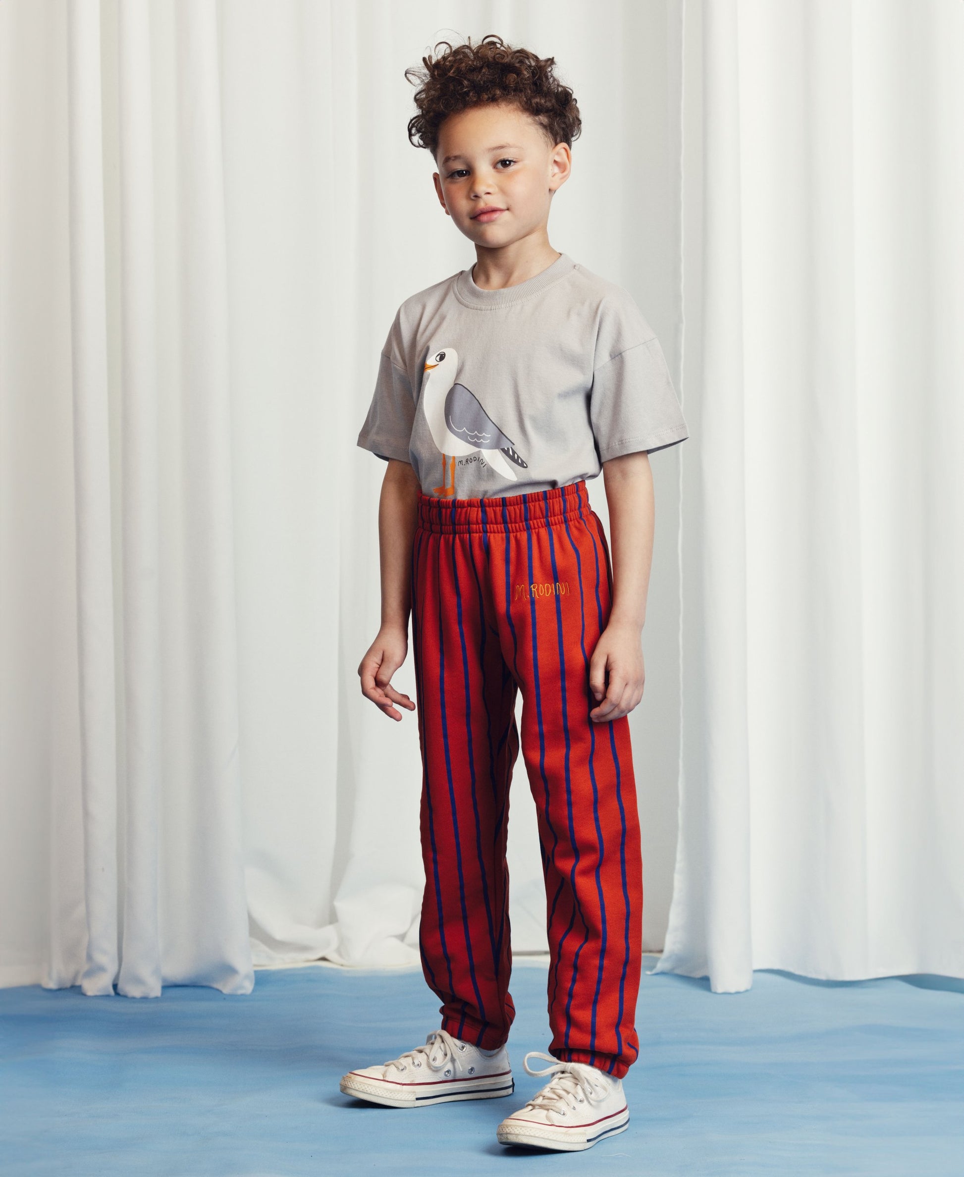 A young child with curly hair wears a grey t-shirt featuring a seagull illustration, paired with red and blue striped trousers. The child stands against a white curtain backdrop on a blue floor, looking confidently at the camera.