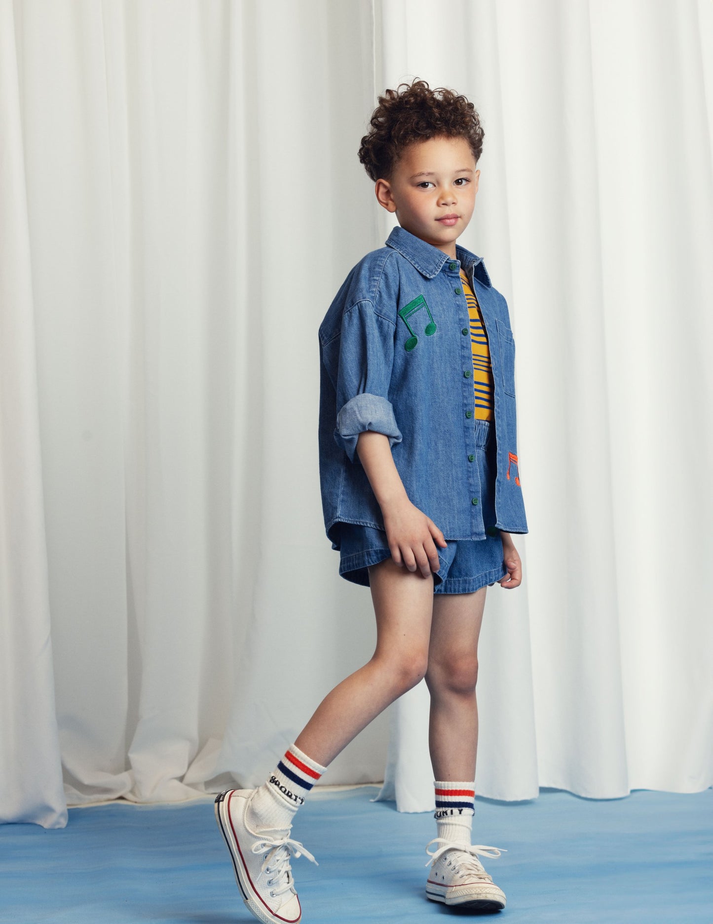 A young child with curly hair wears a denim button-up shirt with embroidered musical notes over a striped top, paired with denim shorts. They complete the look with striped socks and white Converse trainers, posing confidently against a white curtain backdrop.