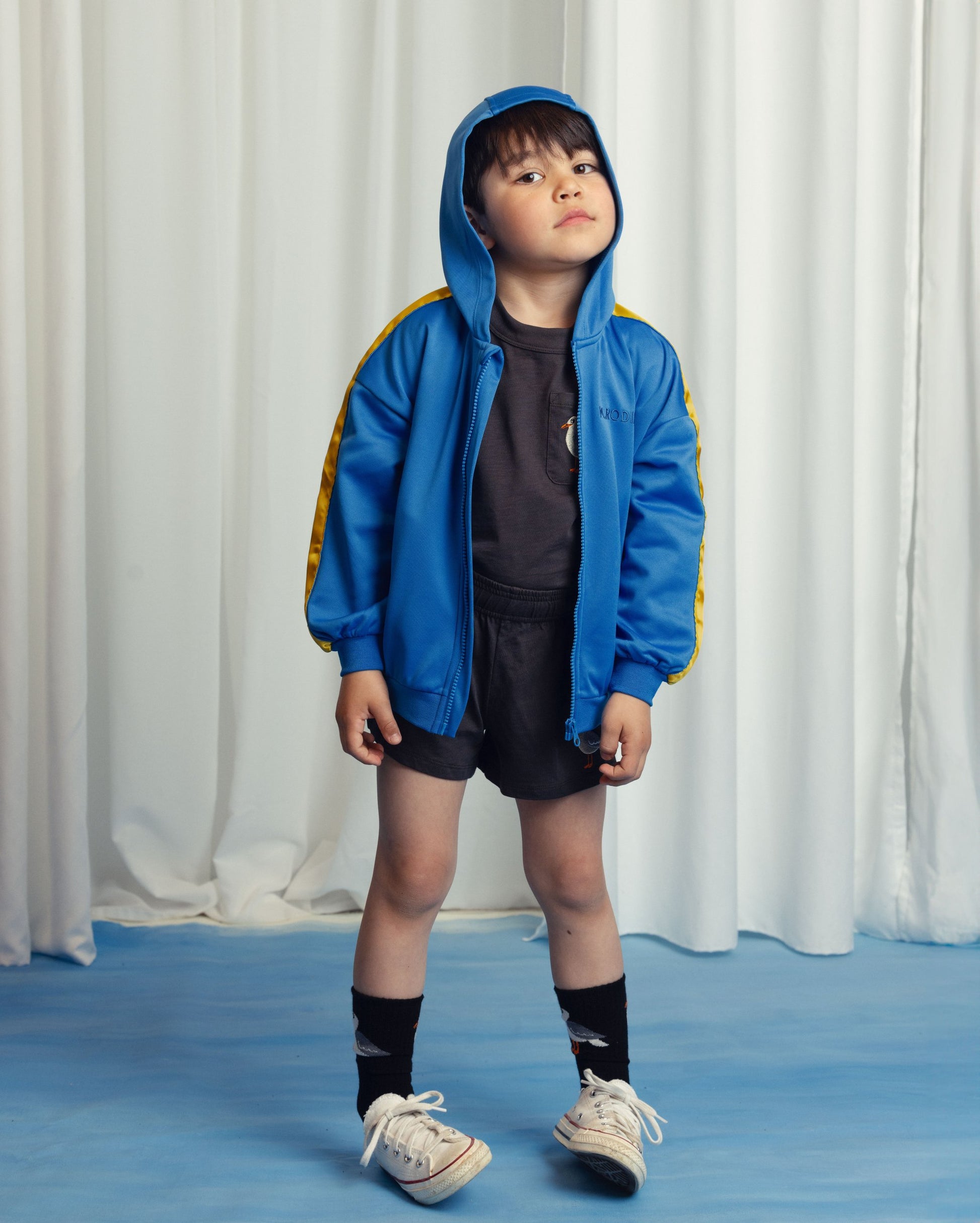 Young boy wearing the Mini Rodini WCT hoodie in bright blue with yellow sleeve stripes, styled with a black t-shirt, black shorts, patterned socks, and white sneakers. He stands against a cream curtain backdrop with a confident expression.