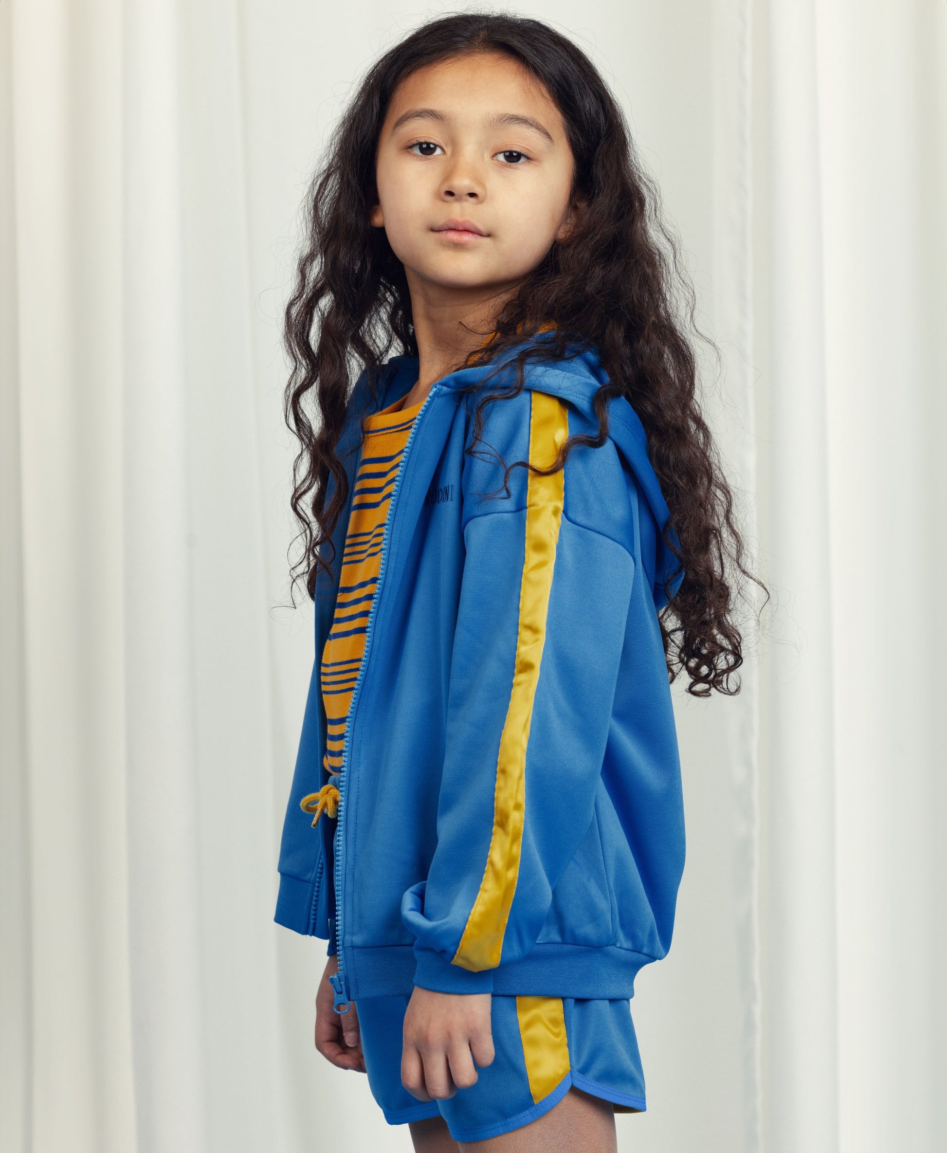 Young girl with long curly hair wearing the Mini Rodini WCT hoodie in blue with yellow sleeve stripes, layered over a yellow and blue striped top and matching blue shorts. She looks directly at the camera with a neutral expression.