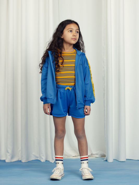 Young girl wearing the Mini Rodini WCT hoodie over a yellow and blue striped top and coordinating blue shorts with a yellow drawstring. She completes the look with white high-top sneakers and striped socks, standing against a minimal cream curtain backdrop.
