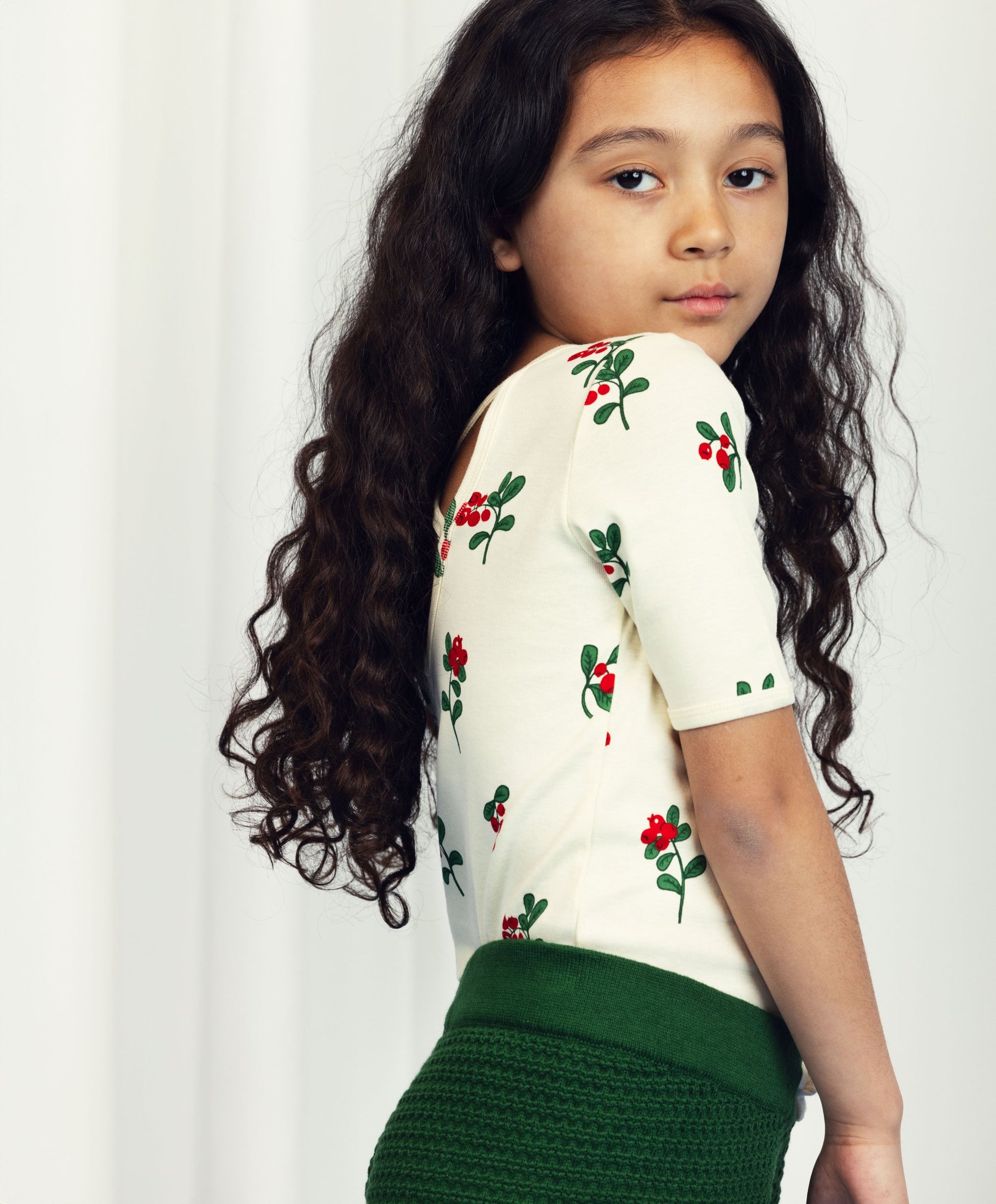 A young girl with long, curly dark hair wears a short-sleeved top with a red berry and green leaf print on an off-white background, tucked into high-waisted, textured green knit shorts. She looks over her shoulder with a calm expression.