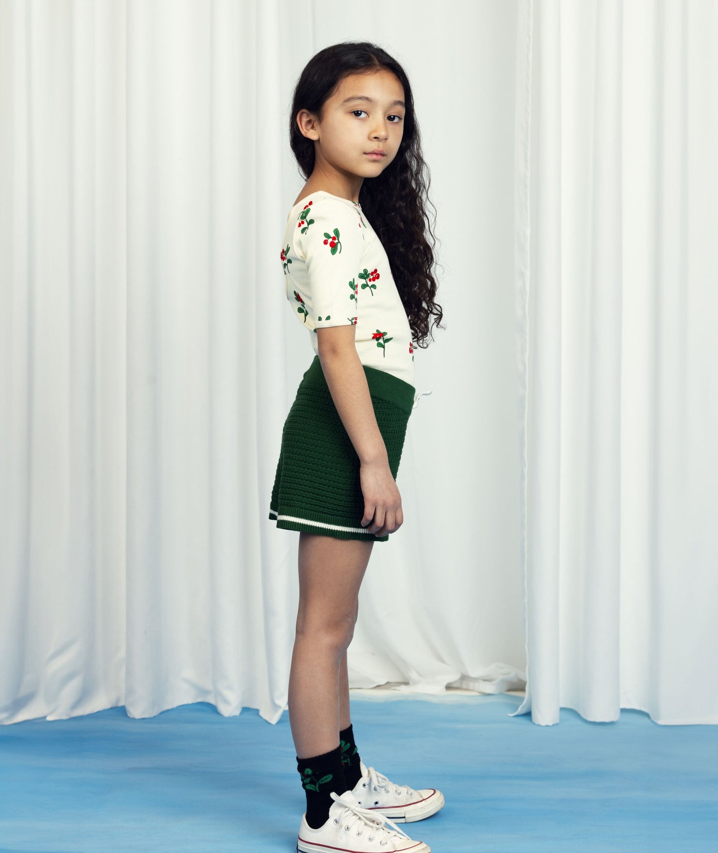 A young girl with long, curly dark hair wears a cream-colored short-sleeved top with a red berry and green leaf pattern, tucked into textured green knit shorts. She pairs the outfit with black socks featuring the same berry pattern and white Converse trainers, standing in profile against a white curtain backdrop.