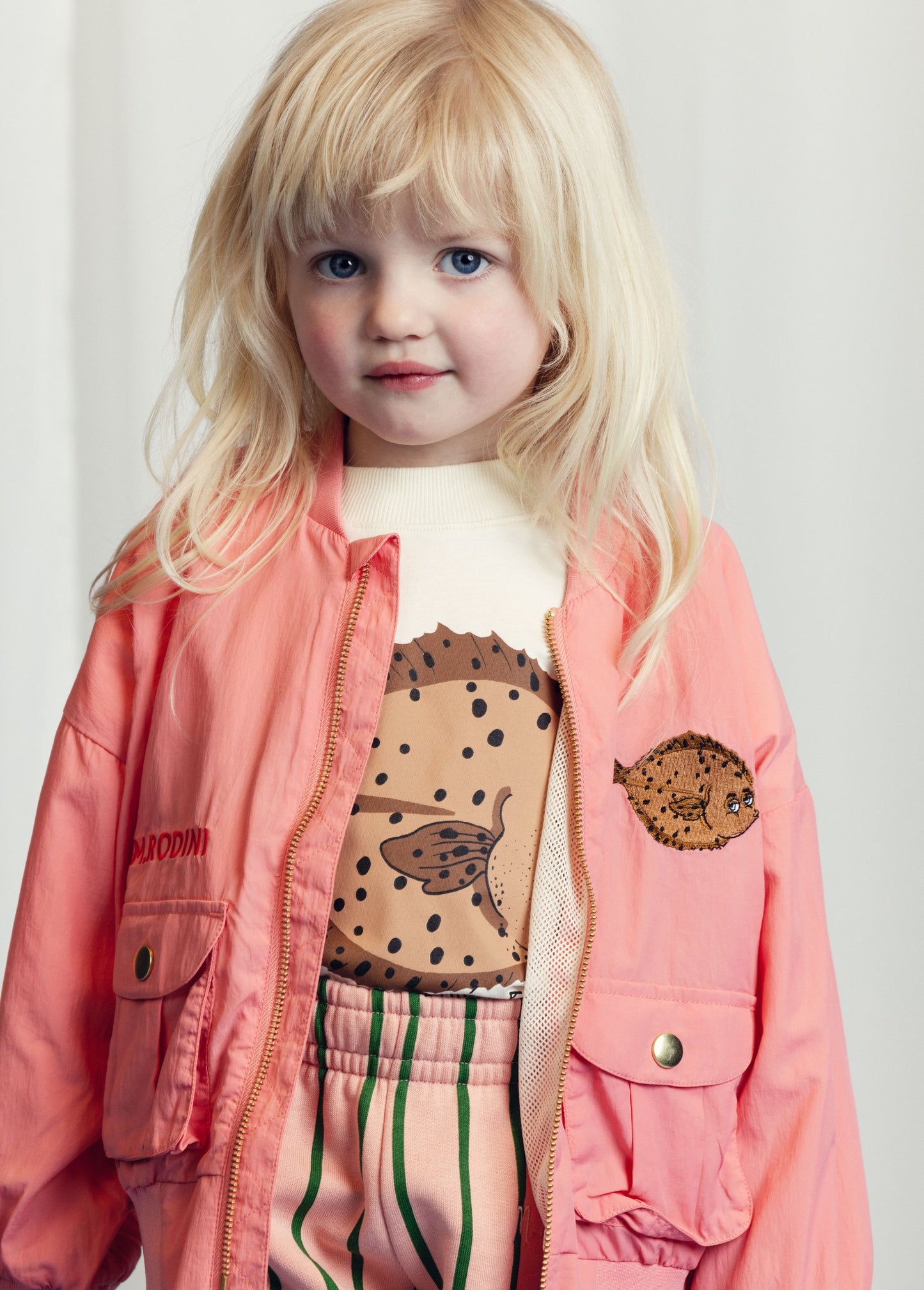 A blonde-haired toddler wears a peach-pink satin jacket adorned with an embroidered pufferfish, layered over a cream top featuring a stingray print. She gazes softly at the camera, with a white curtain backdrop.