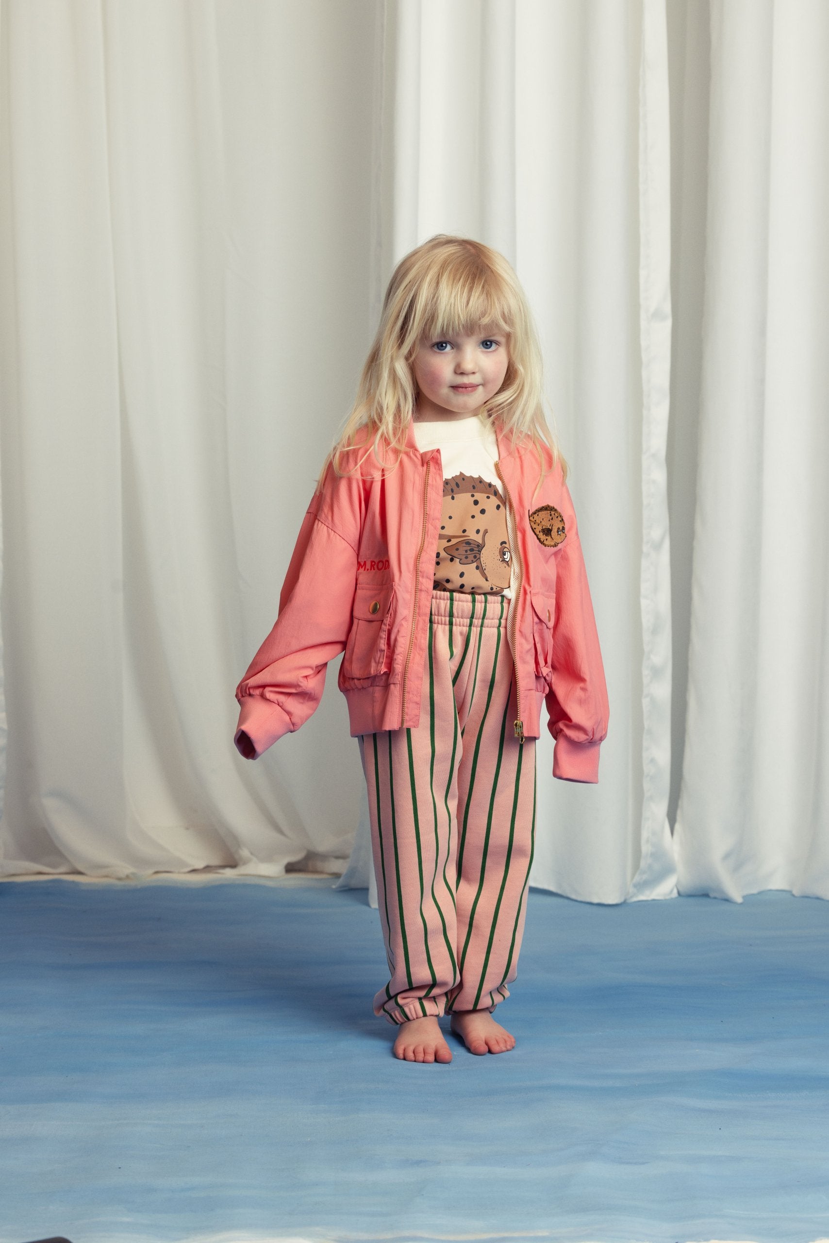 A small child stands barefoot on a blue floor, dressed in a Mini Rodini pink bomber jacket with a pufferfish design and matching striped trousers. They smile subtly in front of a white draped background.