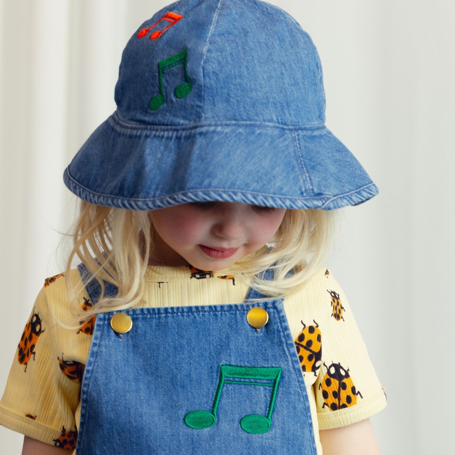 A young child wearing Mini Rodini’s ladybird ribbed T-shirt under denim dungarees with embroidered musical notes, paired with a denim bucket hat.