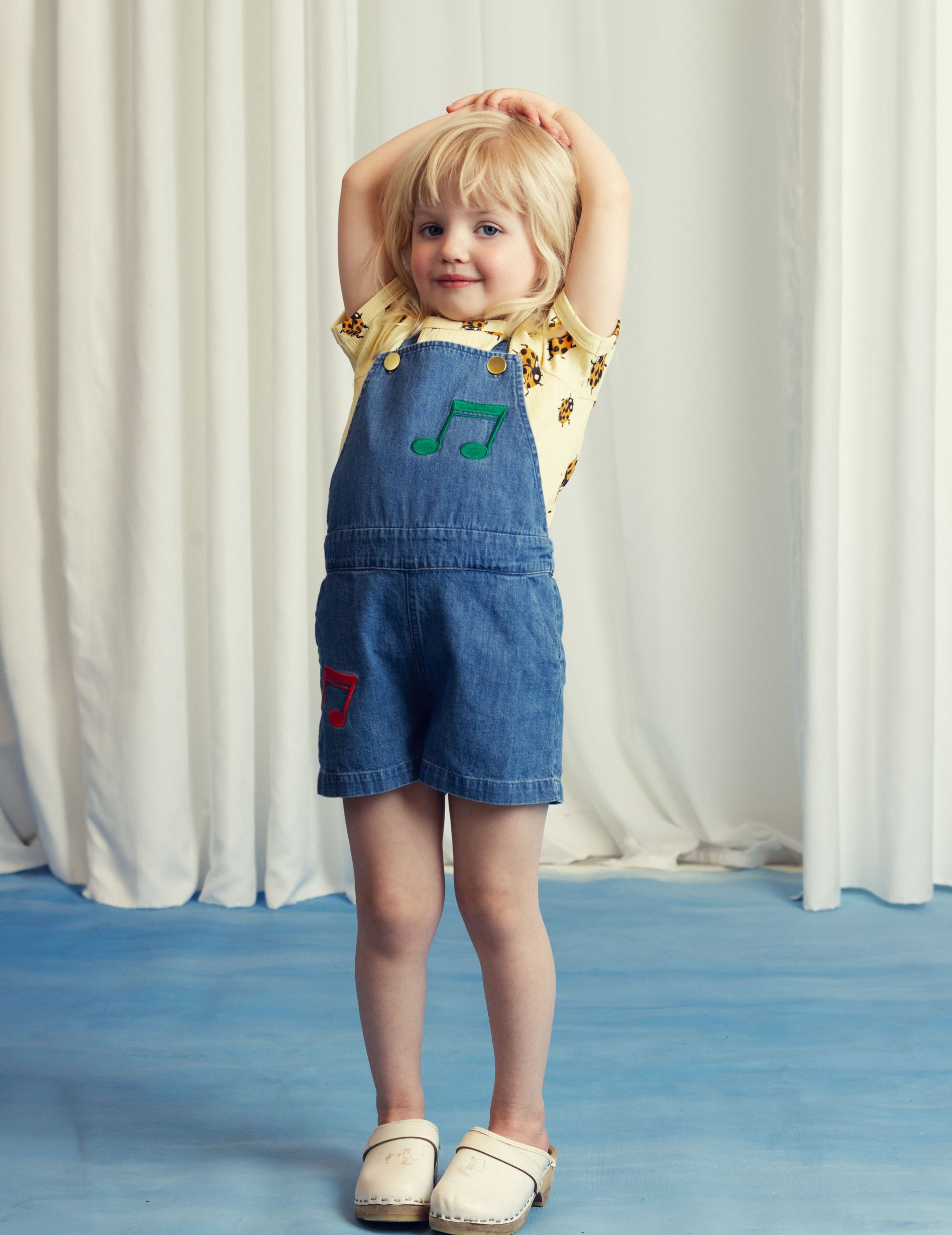 A young child posing in Mini Rodini’s ladybird T-shirt and denim dungaree shorts with embroidered musical notes, styled with wooden clogs.