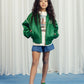 Full-body shot of child wearing jacket: A child poses in the Mini Rodini Note Chenille Satin Baseball Jacket, styled with denim shorts and green clogs. The jacket has a relaxed, slightly oversized fit with a bold, sporty look.