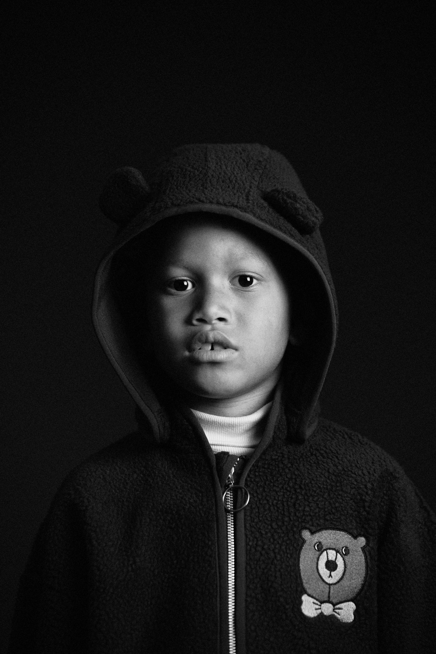 Black and white portrait of a child wearing a bear-themed hoodie against a dark background