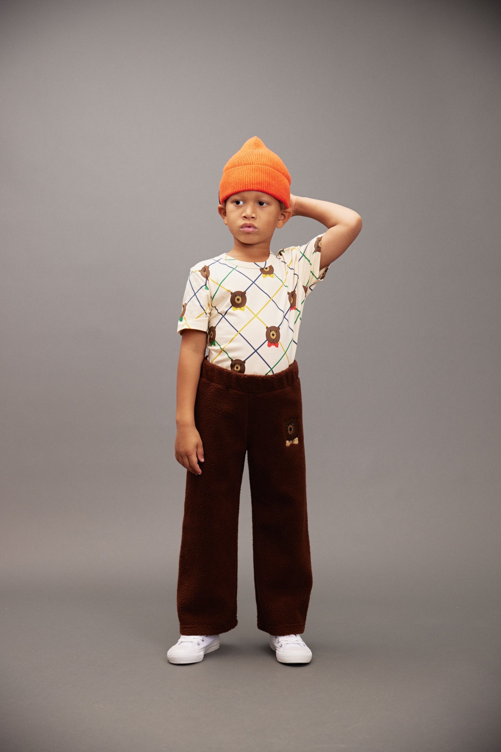 Child wearing an orange beanie, patterned shirt, and brown pants on a gray background