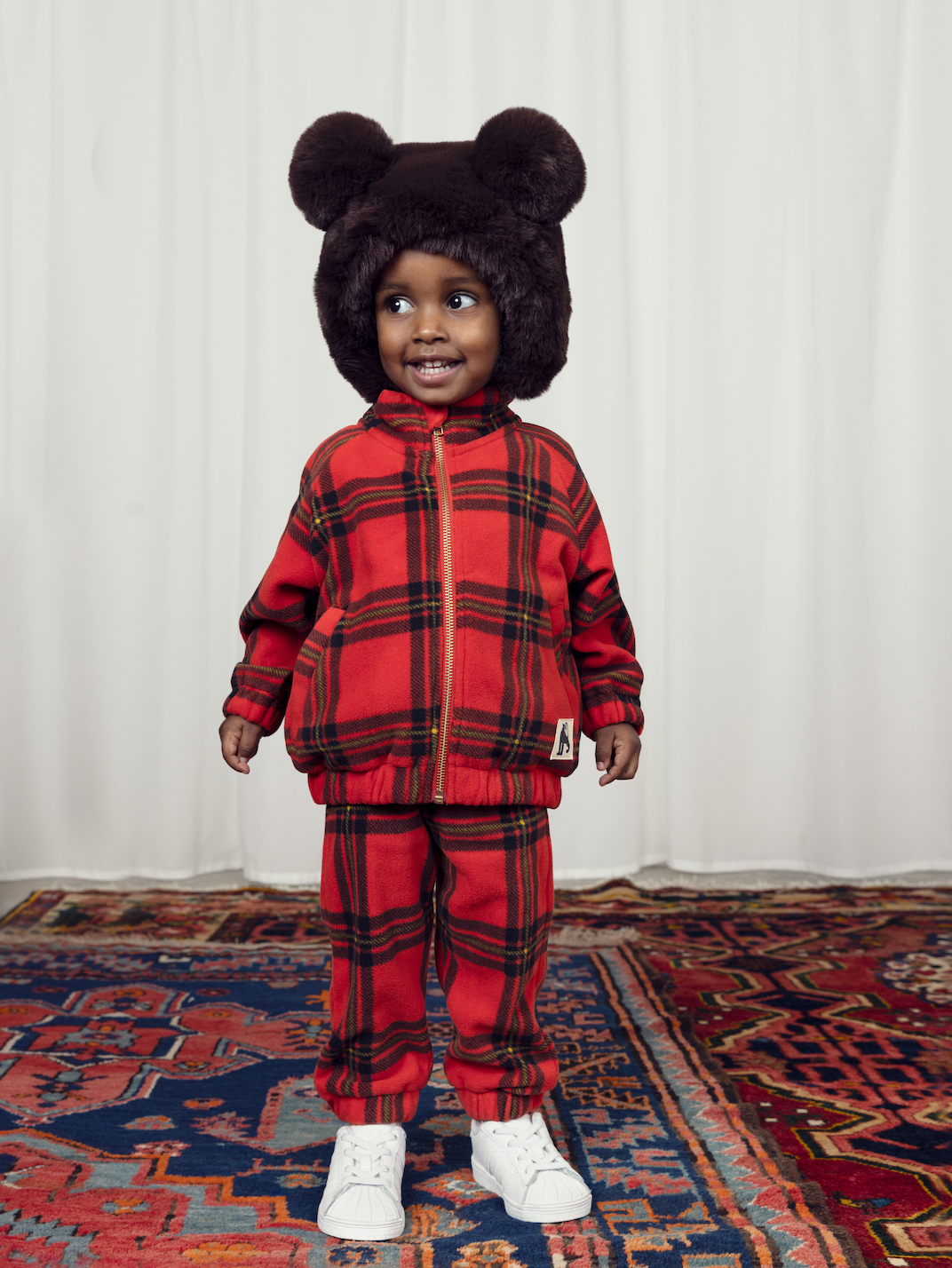 A child in the Mini Rodini children bright red with large check pattern fleece jacket, with a gold plated zip and two side pockets and a high neck