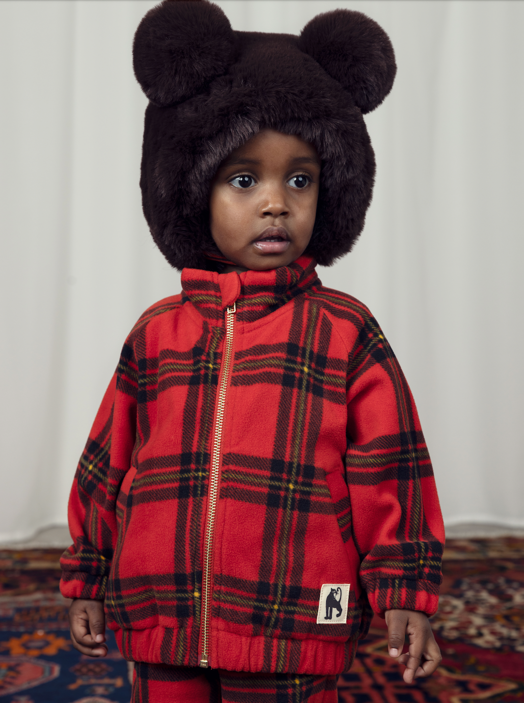 a child in the a Mini Rodini children sbright red with large check pattern fleece jacket, with a gold plated zip and two side pockets and a high neck