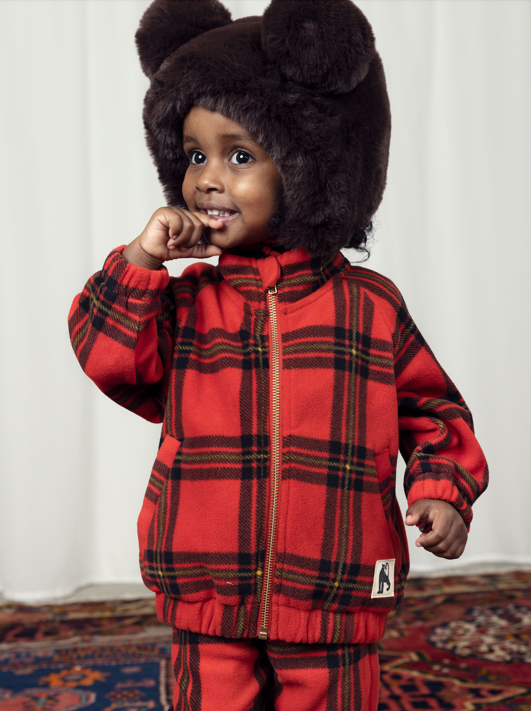 A child in the Mini Rodini children bright red with large check pattern fleece jacket, with a gold plated zip and two side pockets and a high neck