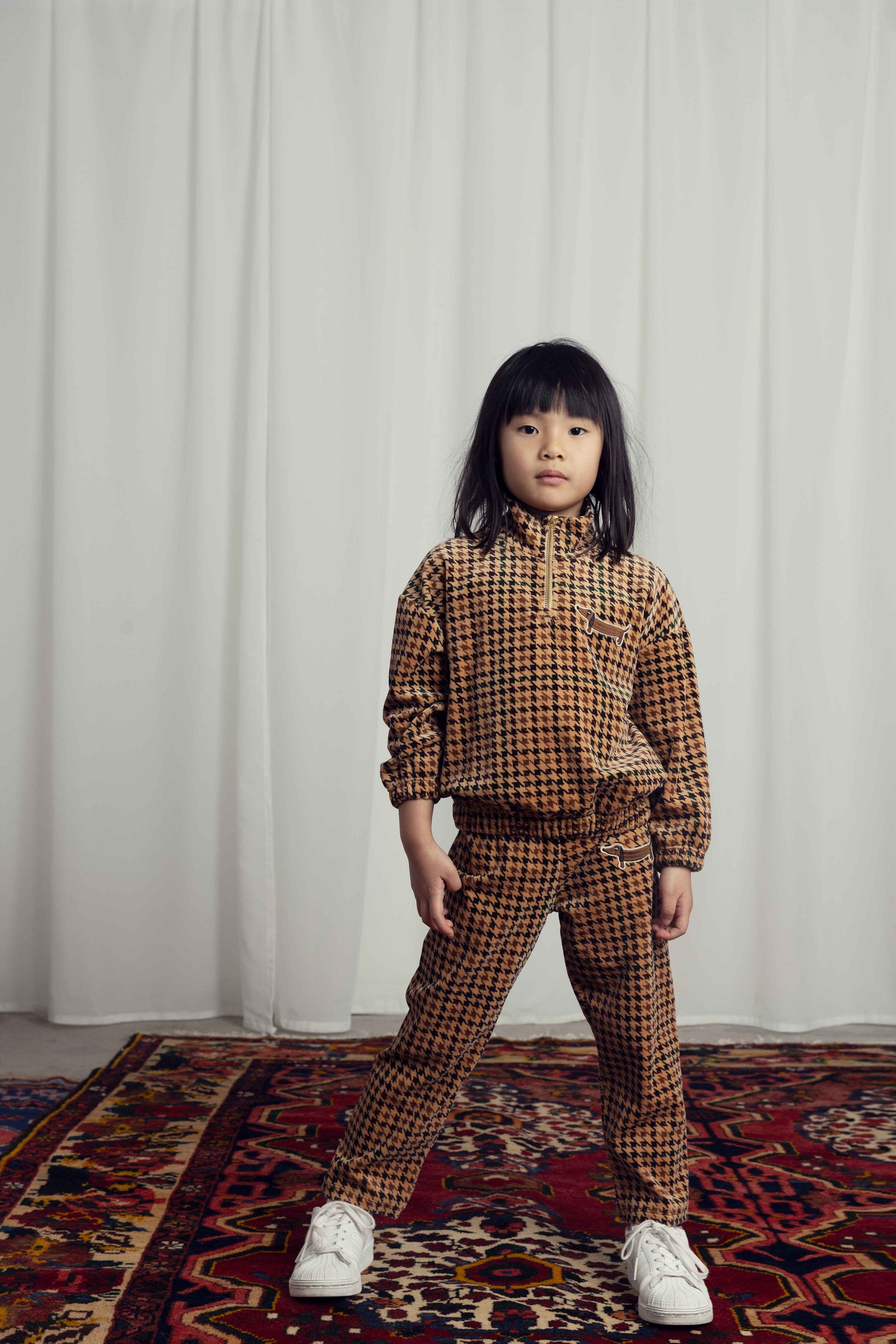 A child in the Mini Rodini houndstooth velour quarter zip tracksuit jacket in a light beige tone with darker brown and black houndstooth all over pattern