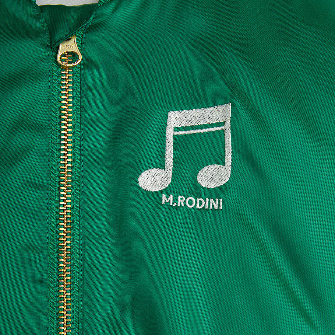 A close-up view of the Mini Rodini Note Chenille Satin Baseball Jacket in vibrant green, featuring an embroidered white musical note and "M. Rodini" text on the chest. The gold YKK zipper adds a sleek finish.