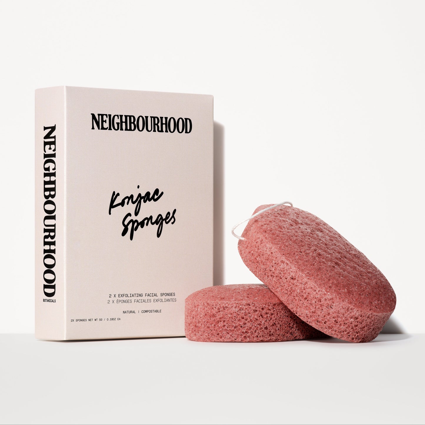 A set of two pink exfoliating konjac sponges with a slightly porous texture, placed next to a minimalist beige box labeled "Neighbourhood Konjac Sponges." The packaging features bold black lettering and states that the sponges are natural and compostable.