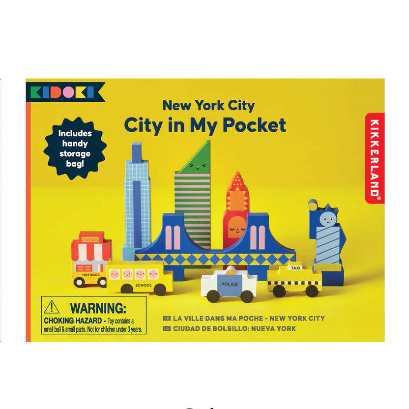 New York City In My Pocket wooden toy set packaging. The box features a yellow background with illustrated wooden blocks showcasing New York landmarks such as the Empire State Building, Statue of Liberty, Brooklyn Bridge, and a yellow taxi. A label highlights the included storage bag. A warning label notes small parts, not suitable for children under 3 years.