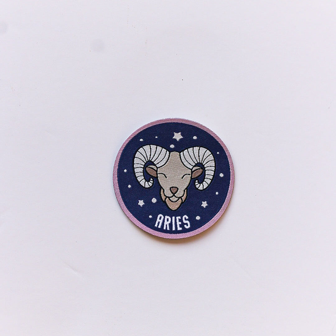 A close-up of an Aries zodiac iron-on patch featuring a stylised ram’s head on a navy background with white stars and a soft pink border.