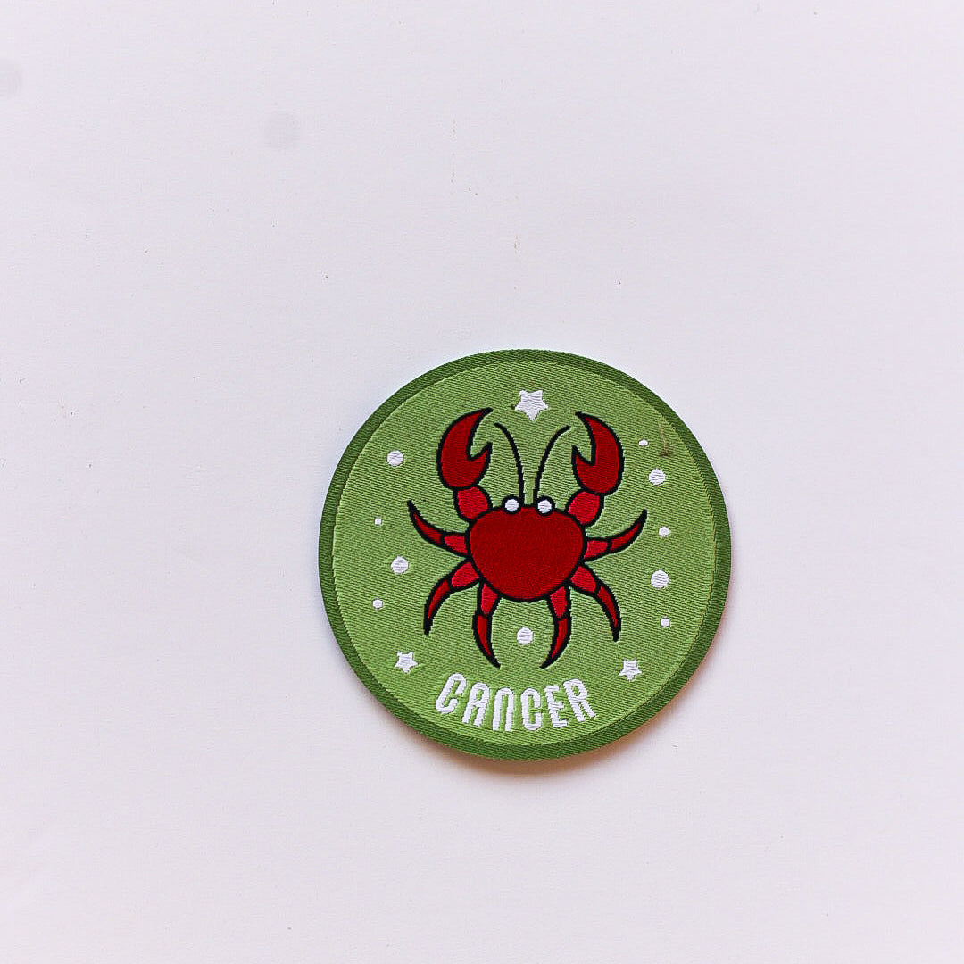 A close-up of a Cancer zodiac iron-on patch featuring a red crab illustration on a green background with white stars and a deep green border.