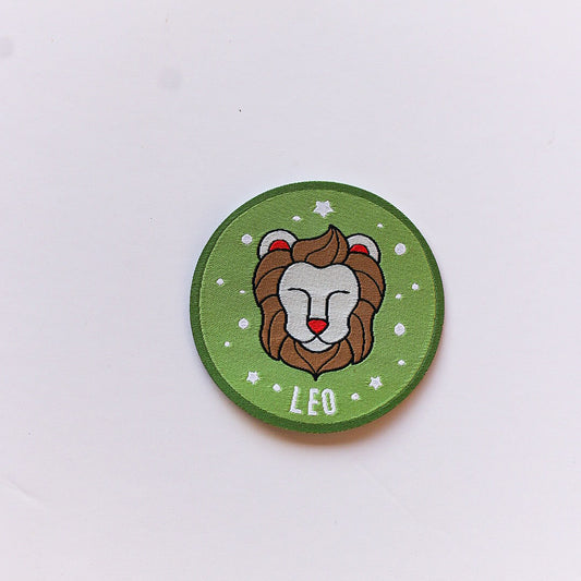 A close-up of a Leo zodiac iron-on patch featuring a lion’s face on a green background with white stars and a soft pink border.