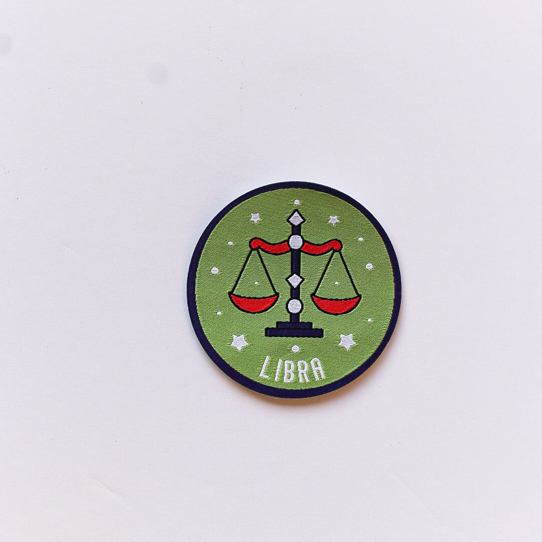 A close-up of a Libra zodiac iron-on patch featuring a set of balanced scales with red details, on a green background with white stars and a dark blue border.