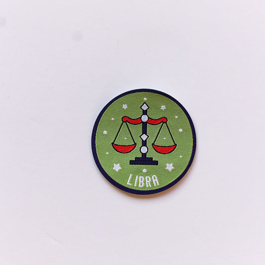 A close-up of a Libra zodiac iron-on patch featuring a set of balanced scales with red details, on a green background with white stars and a dark blue border.