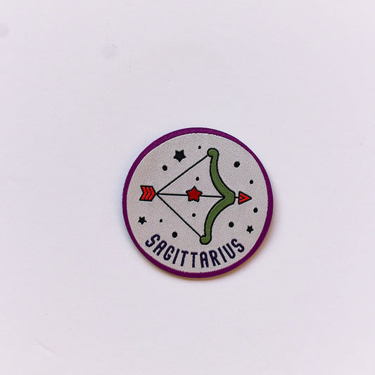 A close-up of a Sagittarius zodiac iron-on patch featuring a green bow and arrow with a red star, set against a cream background with white stars and a deep purple border.