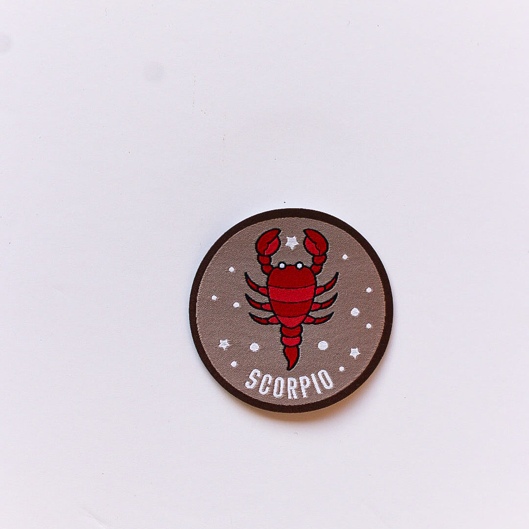 A close-up of a Scorpio zodiac iron-on patch featuring a red scorpion illustration on a brown background with white stars and a dark brown border.
