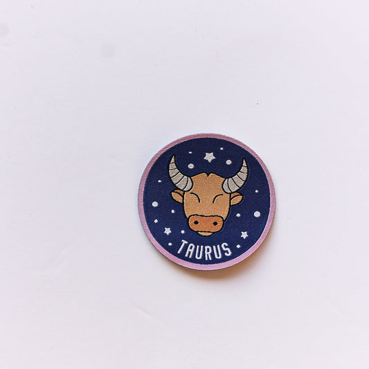 A close-up of a Taurus zodiac iron-on patch featuring a stylised bull’s face on a navy background with white stars and a soft pink border.