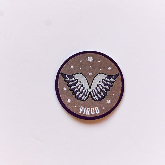 A close-up of a Virgo zodiac iron-on patch featuring a pair of angelic wings on a brown background with white stars and a deep purple border.