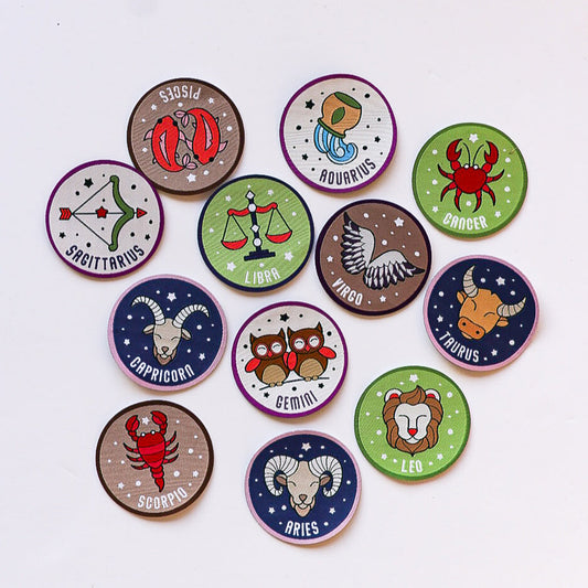 A collection of 12 round zodiac iron-on patches laid out on a white background. Each patch represents a different star sign with a unique illustration, featuring bold colours and playful designs.