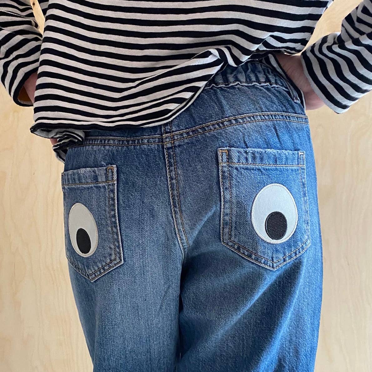 embroidered giant eyeball iron on patches on the back pockets of a blue denim. The person is wearing a black and white stripes full sleeve T-shirt. The iron on patches are black and white with silver border. 